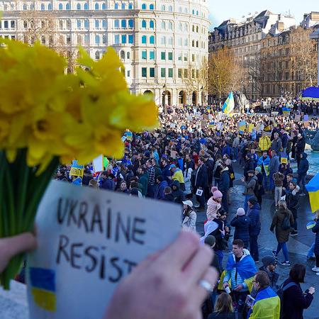 A crowd gathered, holding blue and yellow flags; hands in the foreground hold a sign that says "Ukraine Resist"