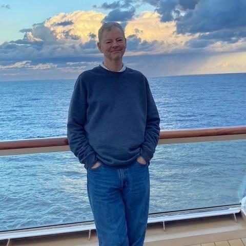Robert Sullivan, with short gray hair and a closed-mouth smile, leaning against a ship's rail with the sea and clouds behind him.