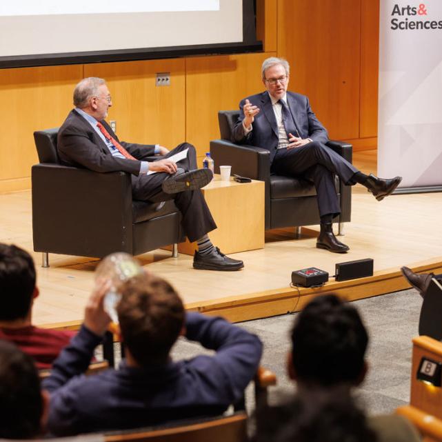 		Two people seated in large chairs, conversing in front of an audience
	