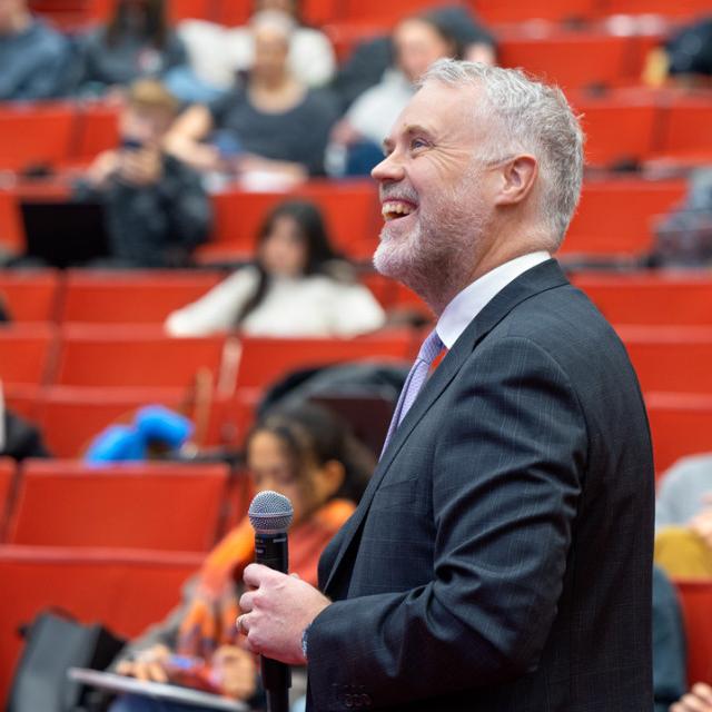 Person in suit holding microphone, in front of a bank of red seats, some occupied