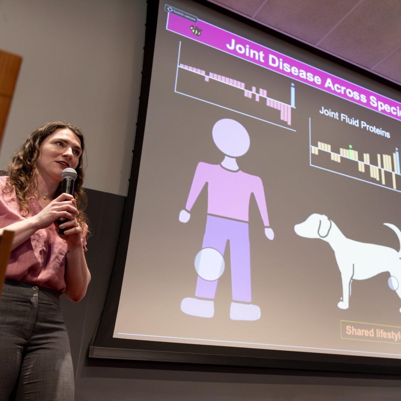 		Person holding a microphone near a large screen projecting a slide about "Joint disease across the species"
	