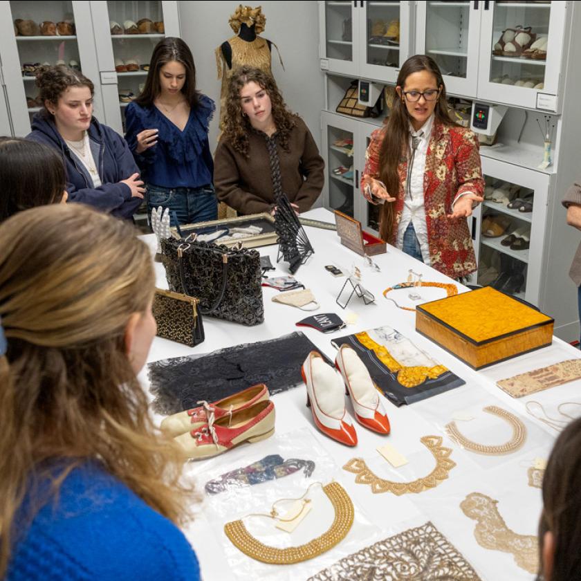 		Several people clustered around a large table, on which shoes, handbags, boxes and collars are displayed
	