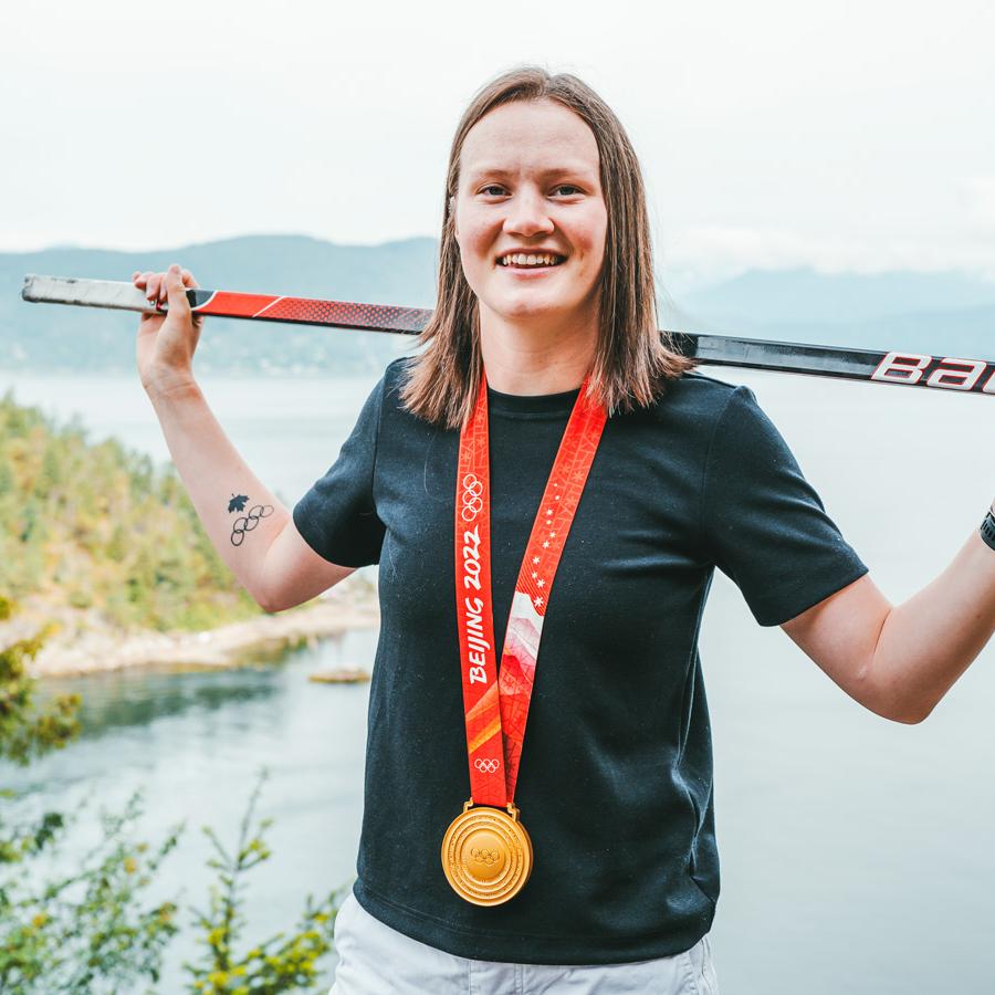 Person standing in front of a body of water, balancing a hockey stick across her back and wearing a gold medal on a red ribbon