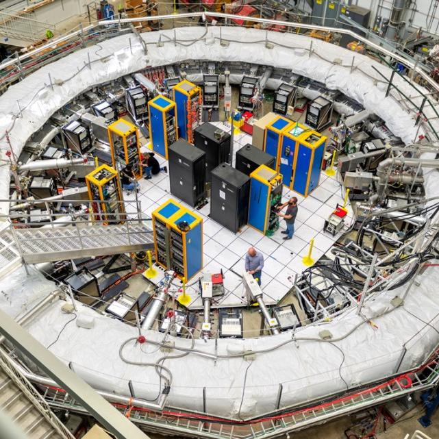 Very large, while ring – a physics experiment setup – dwarfs equipment in its center and people working in it 