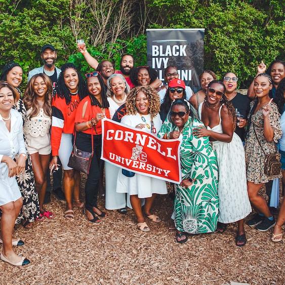 A few dozen people in summer fashions cluster together with a Cornell University banner