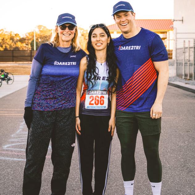 A smiling woman and man in caps and "DARE2TRI" T-shirt stand on either side of a young woman athlete wearing the number "266" on her shirt