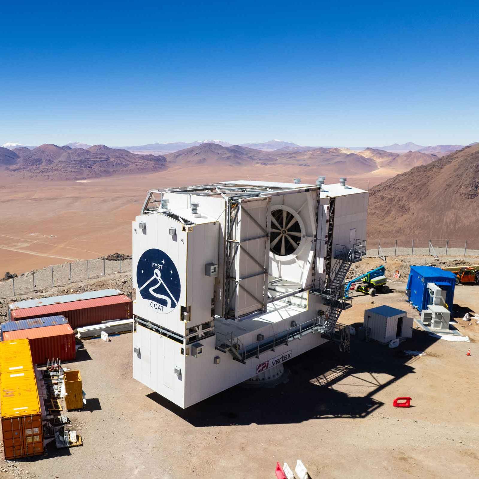 		The telescope, a large white structure with the logo on the front, with construction boxes around it and mountain tops and a plateau in the distance.
	