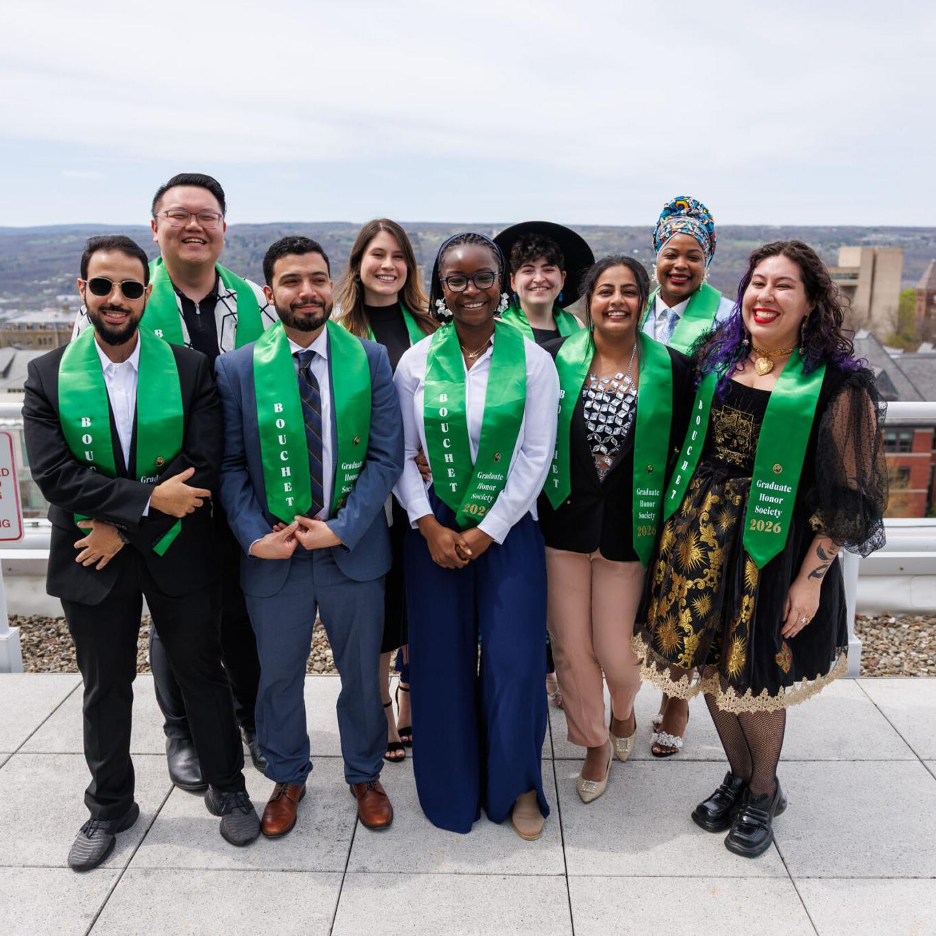 Several people, all wearing green honorary stoles, stand together on a rooftop with a view of a college campus