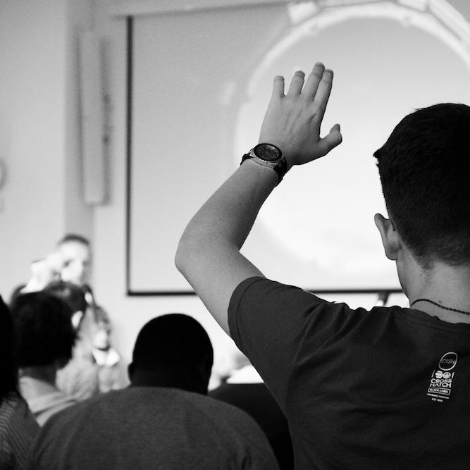Person seen from the back with hand raised in an audience facing a speaker