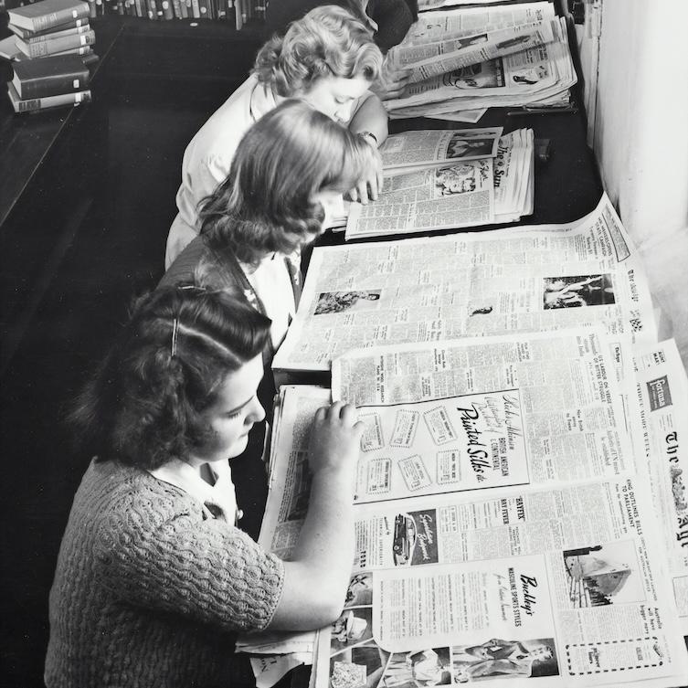 Historic black and white image of five people reading newspapers together at a table