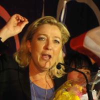 Marine Le Pen with sholuder-length blonde hair and jacket, with hand upraised in the midst of a speech, with French flag in bakcground