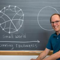 Steven Strogatz in front of a blackboard with "small world" and an illustration on it showing a circle and interconnected lines inside