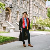 Smiling photo of smiling man with Cornell graduation gown in front of academic builing.