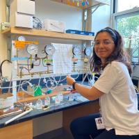 Ligia Coelho, with wire glasses and t-shirt, smiling at the camera next to her lab bench with dials and beakers and wires connecting them