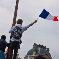 Person, young, holding a French flag in the right hand while holding onto a light post with the left