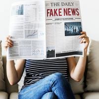  woman on couch holding up newspaper with giant headline that says &quot;Fake News&quot;
