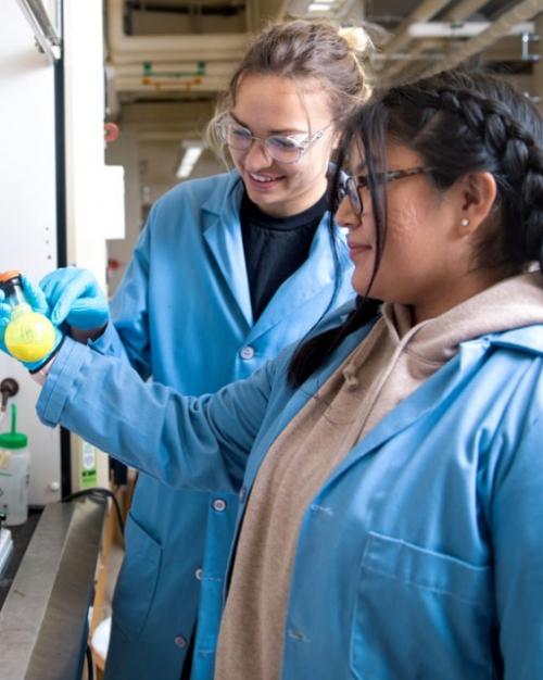 Two students wearing lab coats examining a beaker of something yellow in a lab.