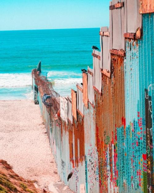A border wall painted different colors blocking a section of beach with the ocean visible. 