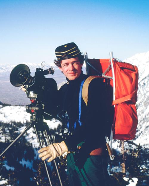 Person in hiking gear with a large camera; mountains in background