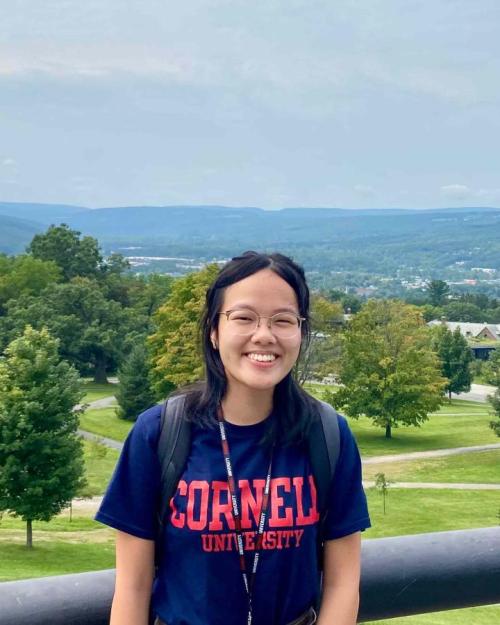 Eunice Ngai in a blue Cornell t-shirt with trees and the Ithaca hills in the background.