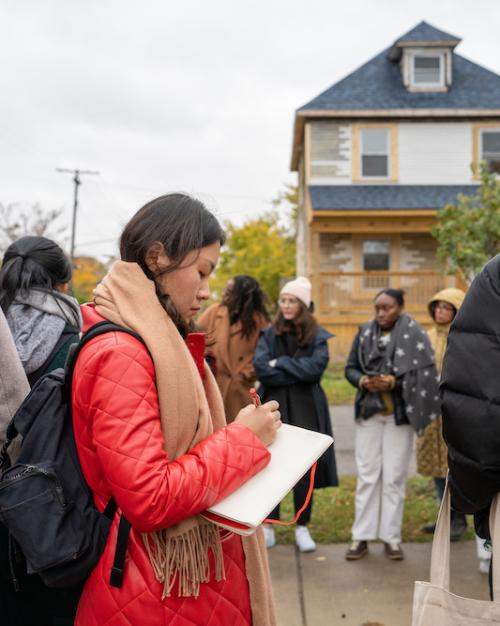 Person wearing a red coat writes in a notebook while standing with a group in front of a row of houses