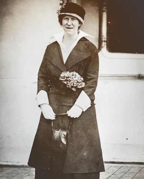 		Black and white photo from 1914: a woman in a dark suit and hat highlighted by flowers stands on a wooden dock
	