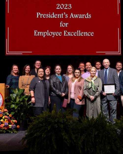 		A few dozen people stand on a stage below a banner: 2023 President's Awards for Employee Excellence
	