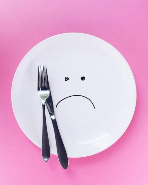 		White plate on a pink background, with a fork and a knife. There is a sad face drawn on the plate
	