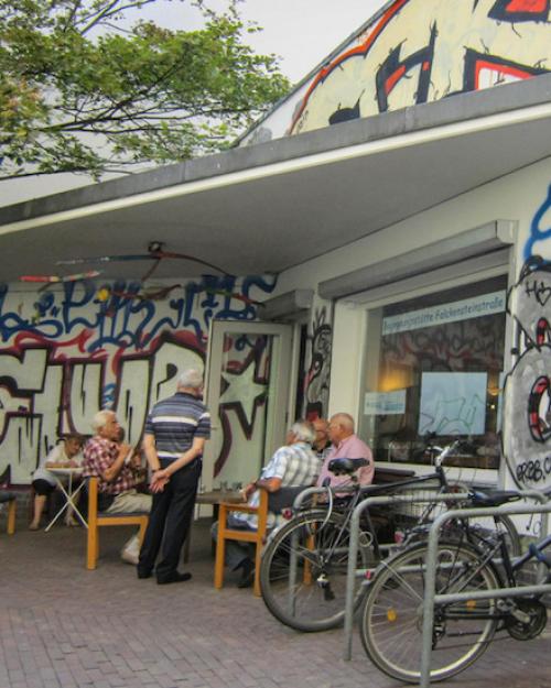 		Elderly people sit in a courtyard near a building with grafiti 
	
