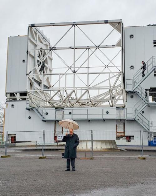 Fred Young in raincoat, holding umbrella, in front of huge metal structure of telescope, with a handful of other people with umbrellas watching him.