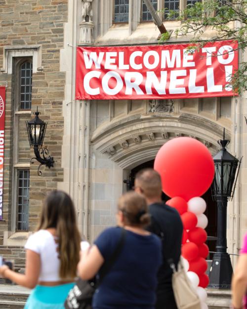 Several people walk past a building with a red and white banner that says "Welcome to Cornell." There are red balloons