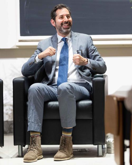 David Folkenflik, with black hair, salt and pepper beard and mustache, in suit and tie, laughing, seated in an armchair.