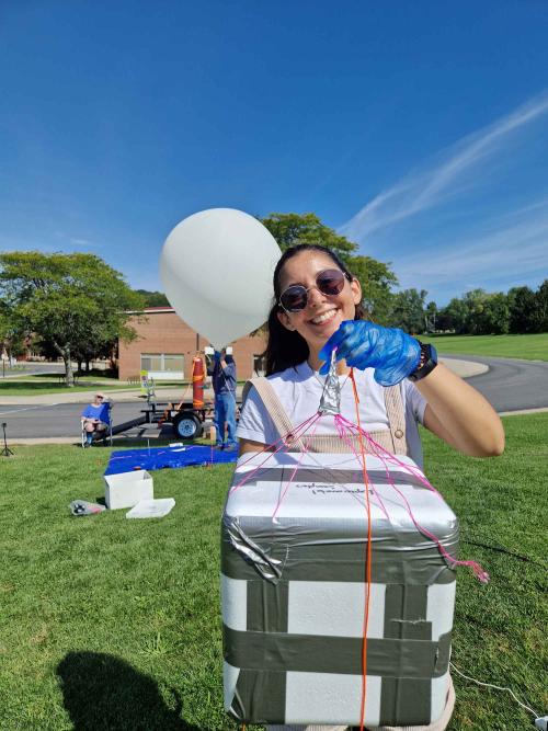 Ligia Coelho, in sunglasses, holding a duct-taped box with string around it next to a big balloon being inflated by a machine.
