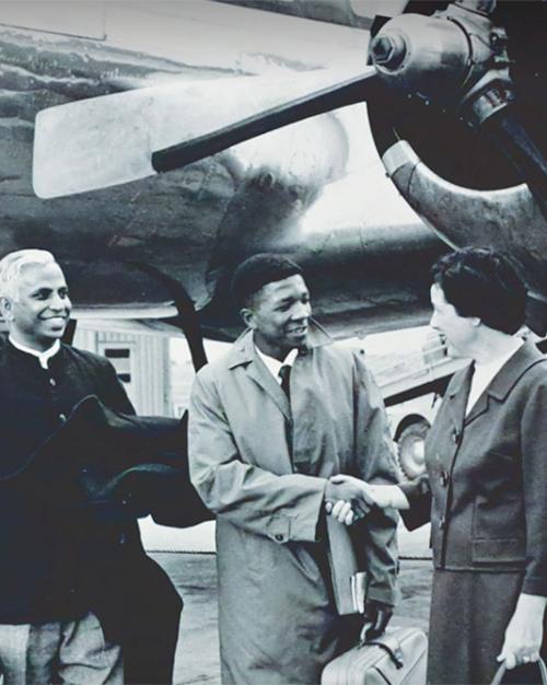 		Black and white historic photo: Five people getting off a plane; one is shaking hands with a person wearing a suit at the bottom of the stairacase
	