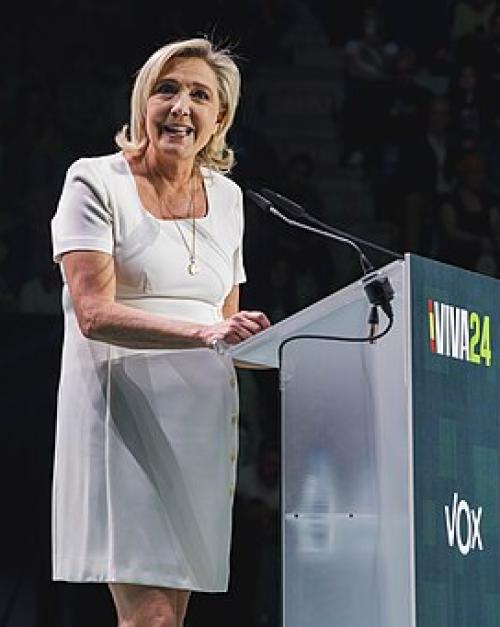 Marine Le Pen in a short white dress facing the audience, standing at a podium that says "Viva24"