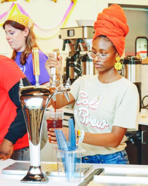 		College students with hats and scarves covering their hair and Saxbys t-shirts make coffee.
	