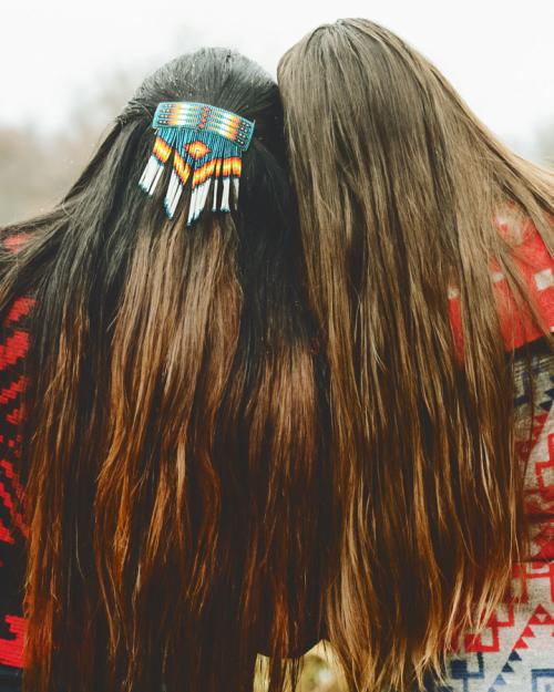 Two women with long hair and wearing Native American-patterned clothing and hairpiece.