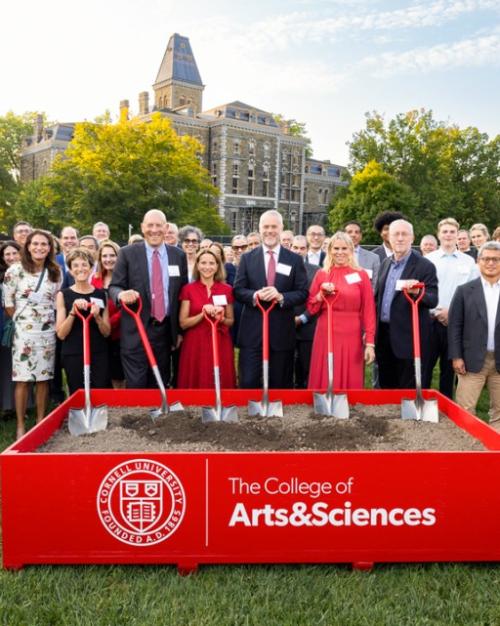 		A crowd of about 75 people stands behind a low box full of dirt; six people in the front hold shovels with red handles during a ceremonial "groundbreaking" event
	