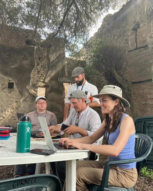 		Four people work at a plastic patio table in the midst of ancient ruins: they are archaeologists on an excavation site
	