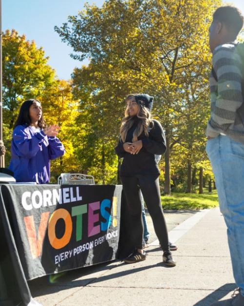 People with backpacks and jeans stand in front of a table, set outdoors, that's labeled "Cornell VOTES"