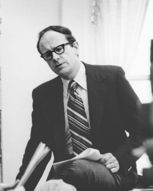 Walter LaFeber in suit and striped tie in his office with bookshelf on one side and a pile of books on the other, talking to someone.