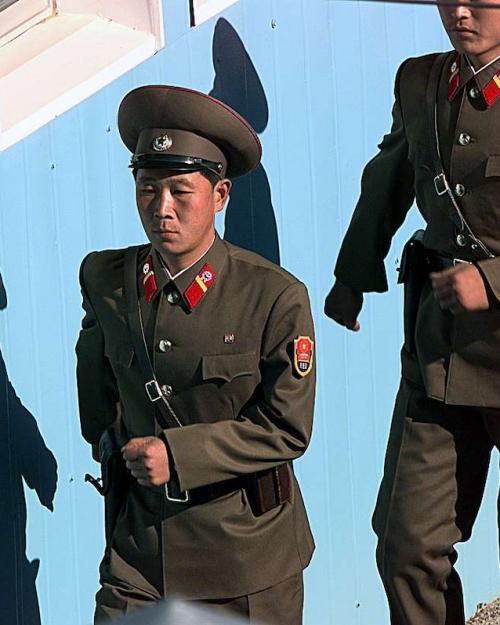 Soldiers in brown formal uniforms march in line beside a blue wall
