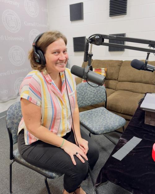 Alexis Boyce, wearing headphones in front of a big microphone, smiles at the camera, wearing a bright colored, striped shirt.