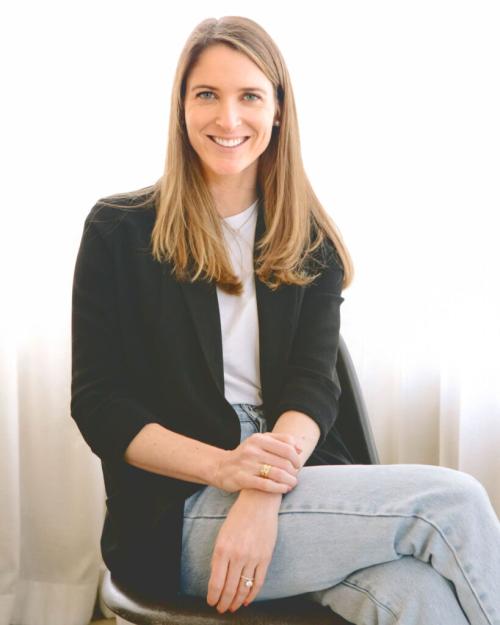 Jenna Barnes, dressed in jeans, open sweater and t-shirt, with long brown hair, seated and smiling.