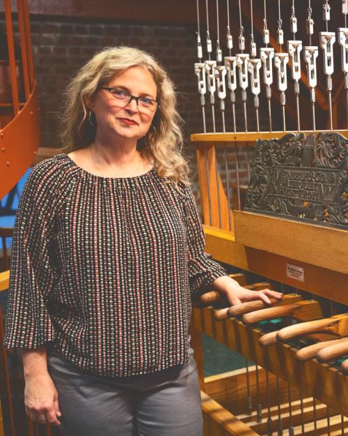 		Jennifer Lory-Moran, in multi-colored stripped shirt and long blond hair, is smiling while standing next to the controls for the Cornell chimes up in McGraw Tower.
	