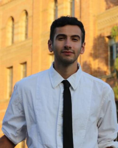 Jeremy Peschard Pórtela with white shirt and black tie, a faint beard and mustache, in front of a sunlight building.