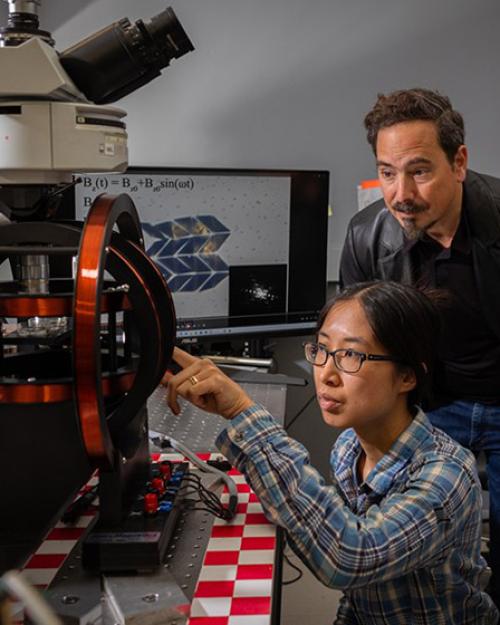 		Three people look intensely at a small black and red machine in a science laboratory
	