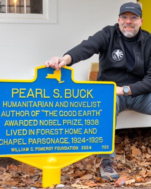 Person sits on a porch with one hand on a blue historic marker