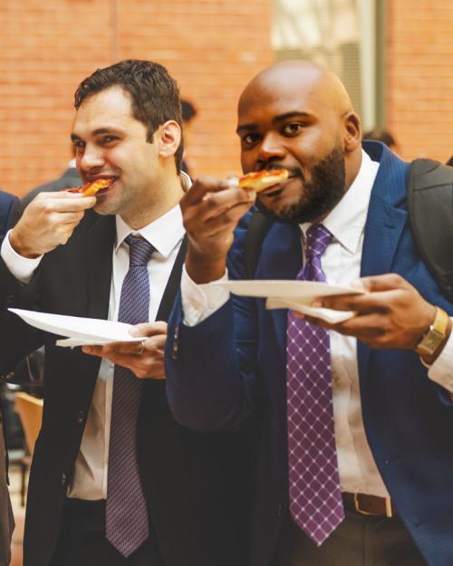 Four people wearing suits, all chomping on slices of pizza and holding plates