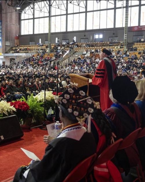 Large indoor gymnasium (Cornell's Barton Hall) decorated with pointsettias and filled with people wearing caps and gowns